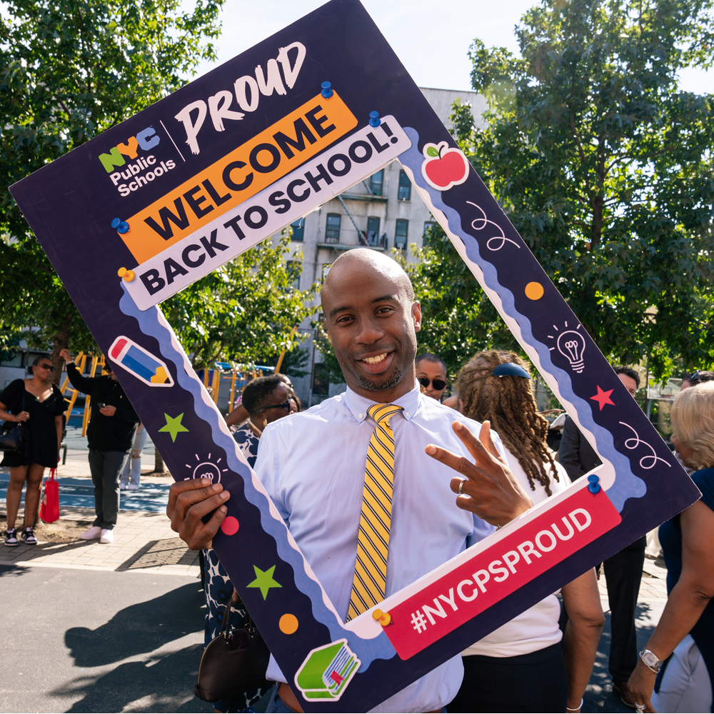 Chancellor Samuels smiling while holding a giant frame around his upper torse that reads, "NYC Public Schools Proud, welcome back to school, #NYCPSProud'