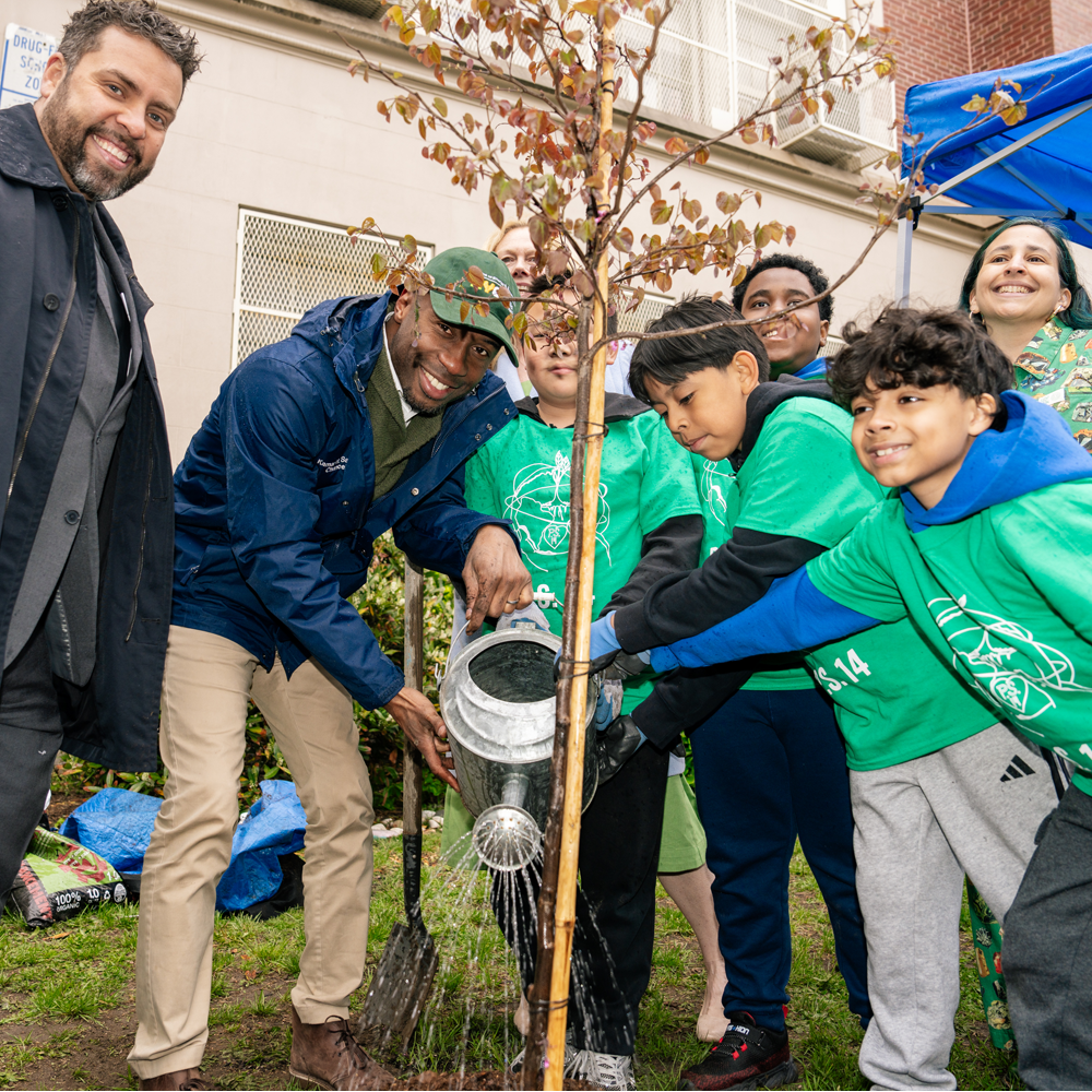 Group of PS 14 students and community members standing with Chancellor Samuels as they water a newly planted tree sapling together
