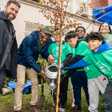 Group of PS 14 students and community members standing with Chancellor Samuels as they water a newly planted tree sapling together