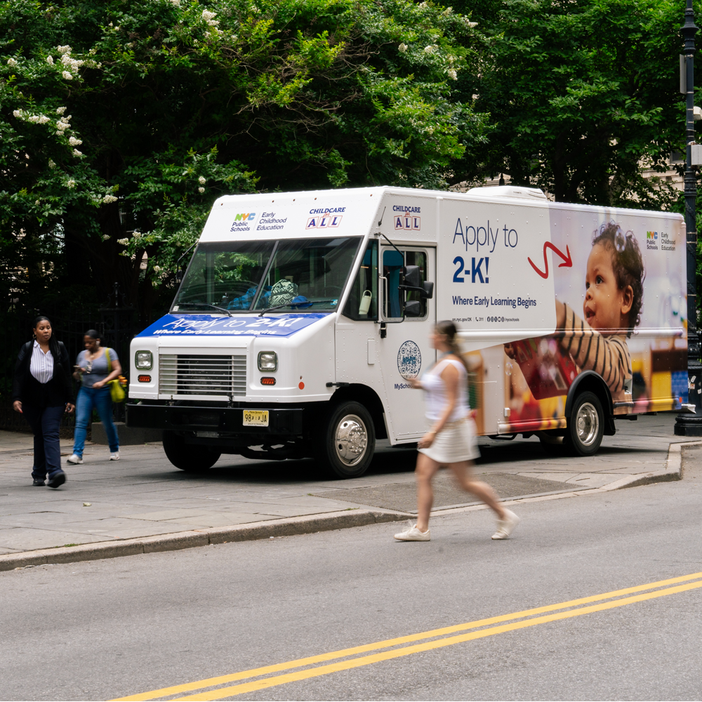 Side view of a large van with signage for NYCPS' Childcare for All program. 'Apply to 2-K' is spelled out in large letters alongside an image of a young child.