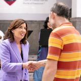 Governor Kathy Hochul (left) shaking a man's hand during her visit to one of NYC's high schools in July 2025 to discuss the upcoming Cell Phone and Electronic Device ban for NYS public schools.