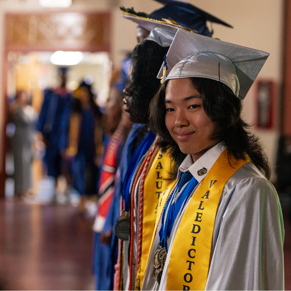 Side view of a young adult wearing a light blue graduation cap and gown featuring a sash that reads, 'Valedictorian.'