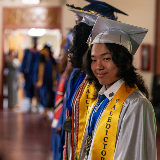 Side view of a young adult wearing a light blue graduation cap and gown featuring a sash that reads, 'Valedictorian.'