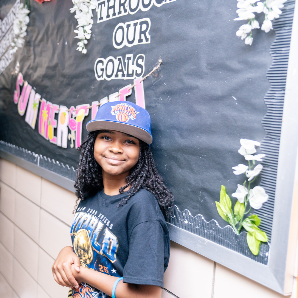 An elementary school-aged student leaning against a school hallway wall and wearing a Knicks hat and a Knicks-themed championship t-shirt.