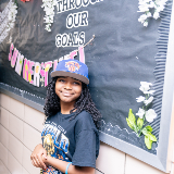 An elementary school-aged student leaning against a school hallway wall and wearing a Knicks hat and a Knicks-themed championship t-shirt.