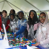 Six students stand at a blue table to work on a sustainability activity. They are wearing rain gear and ponchos.