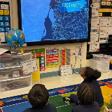 Three students sit on the floor of a classroom looking at a screen that shows a map of New York City's Waterways with the title 'Freshwater' displayed in blue.