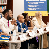 Side view of four District 10 stakeholders, sitting together at a table, listening to the Chancellor and the Governor provide details about the upcoming cell phone policy.