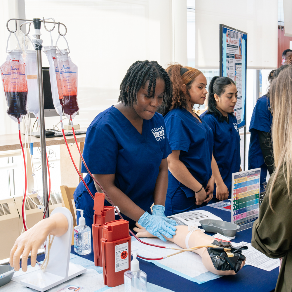 A teenager wearing a nursing uniform leaning over a prosthetic arm and seeking a vein for a nearby blood IV bag.