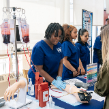 A teenager wearing a nursing uniform leaning over a prosthetic arm and seeking a vein for a nearby blood IV bag.