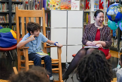 An X036 student sitting in a chair next to Chancellor Melissa Aviles-Ramos, who is seated in front of a library full of students.