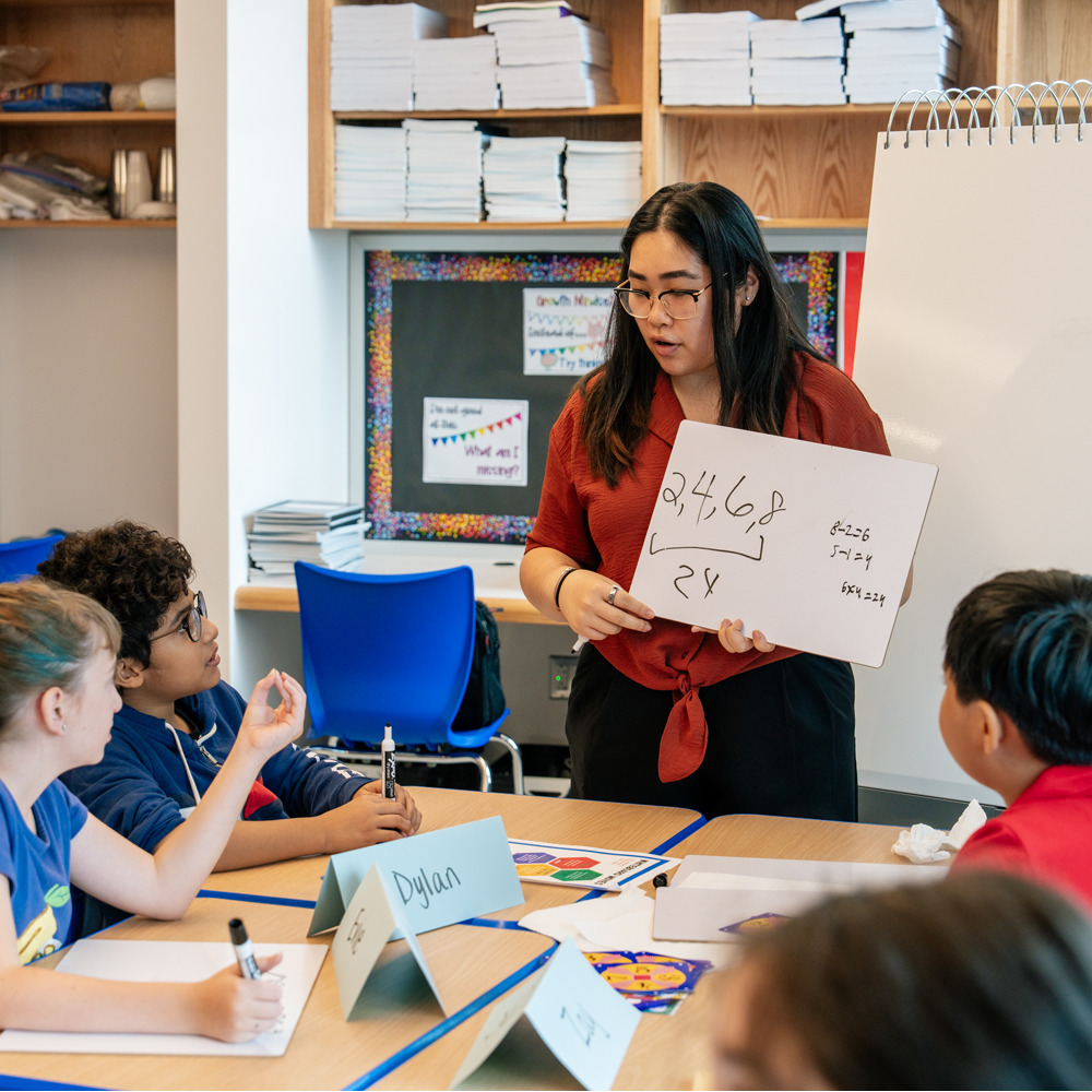 Teacher holding a dry erase board while gesturing to nearby students to answer her written number pattern.