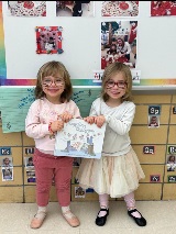 Two young girls standing next to one another while holding a book called "Angelina Ballerina" together. Both girls have cat-like facepaint.