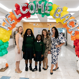 Five women, including two elementary school-aged students (green uniforms, center left) and Chancellor Melissa Aviles-Ramos, posing together underneath an archway of balloons and a message, 'Welcome Back.'