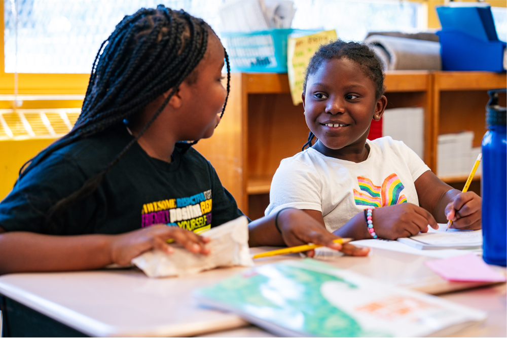 Close-up view of two young elementary-school aged girls as they are sitting at their desks and interacting with each other while inside a classroom.