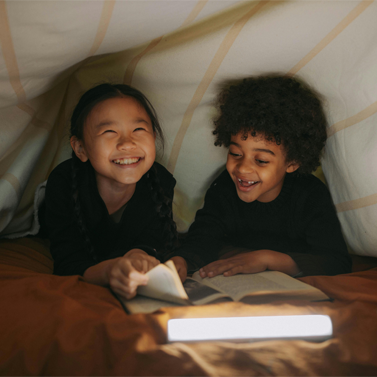 Close-up of two elementary-school aged children, a boy and a girl, reading a book together inside of a makeshift, homemade tent.