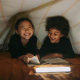 Close-up of two elementary-school aged children, a boy and a girl, reading a book together inside of a makeshift, homemade tent.