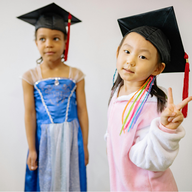 Two young girls wearing graduation caps. One of the girls is holding up two fingers in a 'V.'