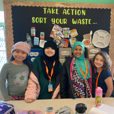 Four students stand in front of a bulletin board that reads 'Take Action, Sort Your Waste,' and explains how to properly sort your trash, recycling, and compost.