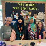 Four students stand in front of a bulletin board that reads 'Take Action, Sort Your Waste,' and explains how to properly sort your trash, recycling, and compost.