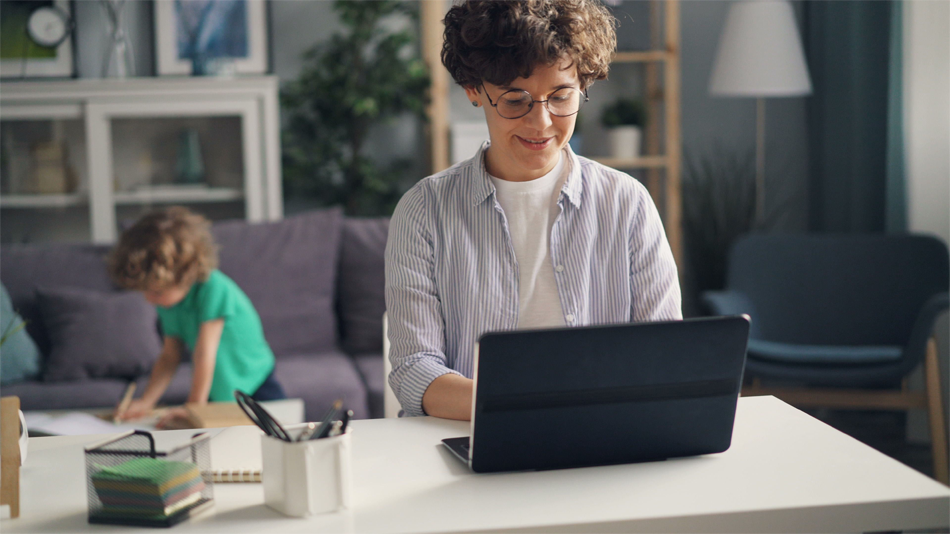 A woman in the foreground of a living room scene working on a laptop with a smile on her face while a child can be seen behind her standing at a coffee table and holding a pencil.