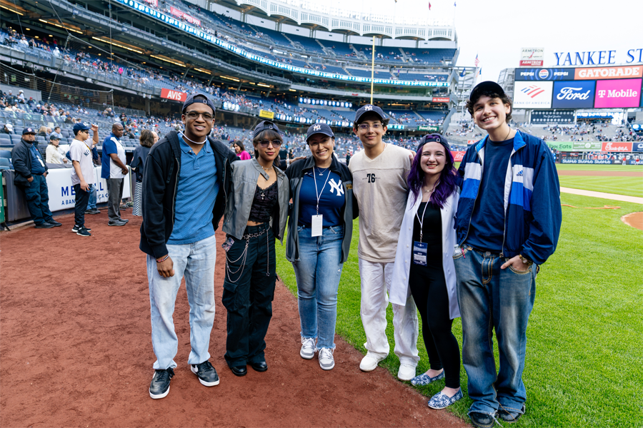 All five of 2025's Yankees Stonewall Scholarship winners standing near home plate at Yankee Stadium with Chancellor Melissa Aviles-Ramos in the middle with a Yankees t-shirt and hat.