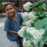 Square crop of young girl standing next to a bush featuring white cherry blossoms.