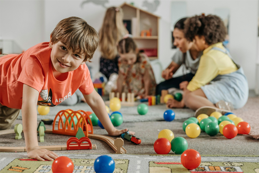 A young boy smiling at the viewer while crawling over a play mat area inside of a classroom that's covered in colored balls and wooden train tracks.