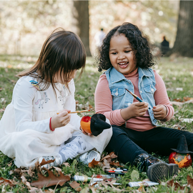 Two young girls sitting on grass while painting and designing pumpkins with witch-inspired paraphernalia.