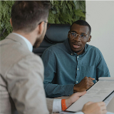 Photo featuring a young man sitting in an office chair and facing an older, suited man in what seems like an interview or meeting of some kind.